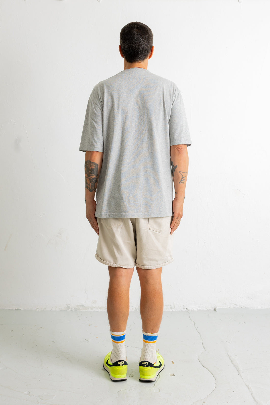 A man viewed from behind wearing a light gray short-sleeve t-shirt, beige shorts, white socks with blue and orange stripes, and neon yellow sneakers, standing against a white wall.